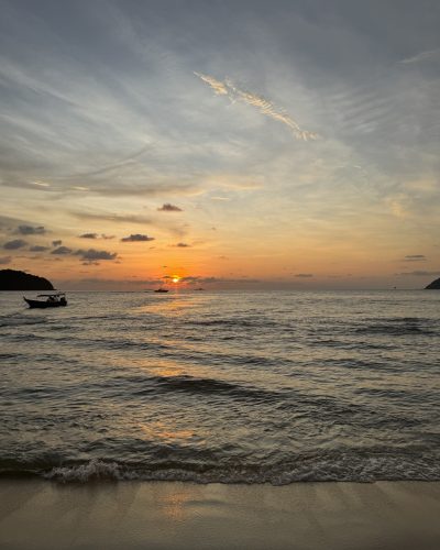 Sonnenuntergang an einem Strand auf Langkawi in Malaysia mit ruhigem Meer, kleinen Booten und orangefarbenem Licht am Horizont.