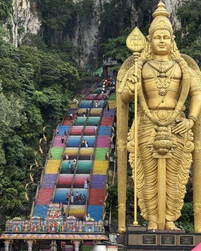 Goldene Murugan-Statue vor den bunten Treppen der Batu Caves in Malaysia mit Kalksteinfelsen und tropischer Vegetation im Hintergrund.