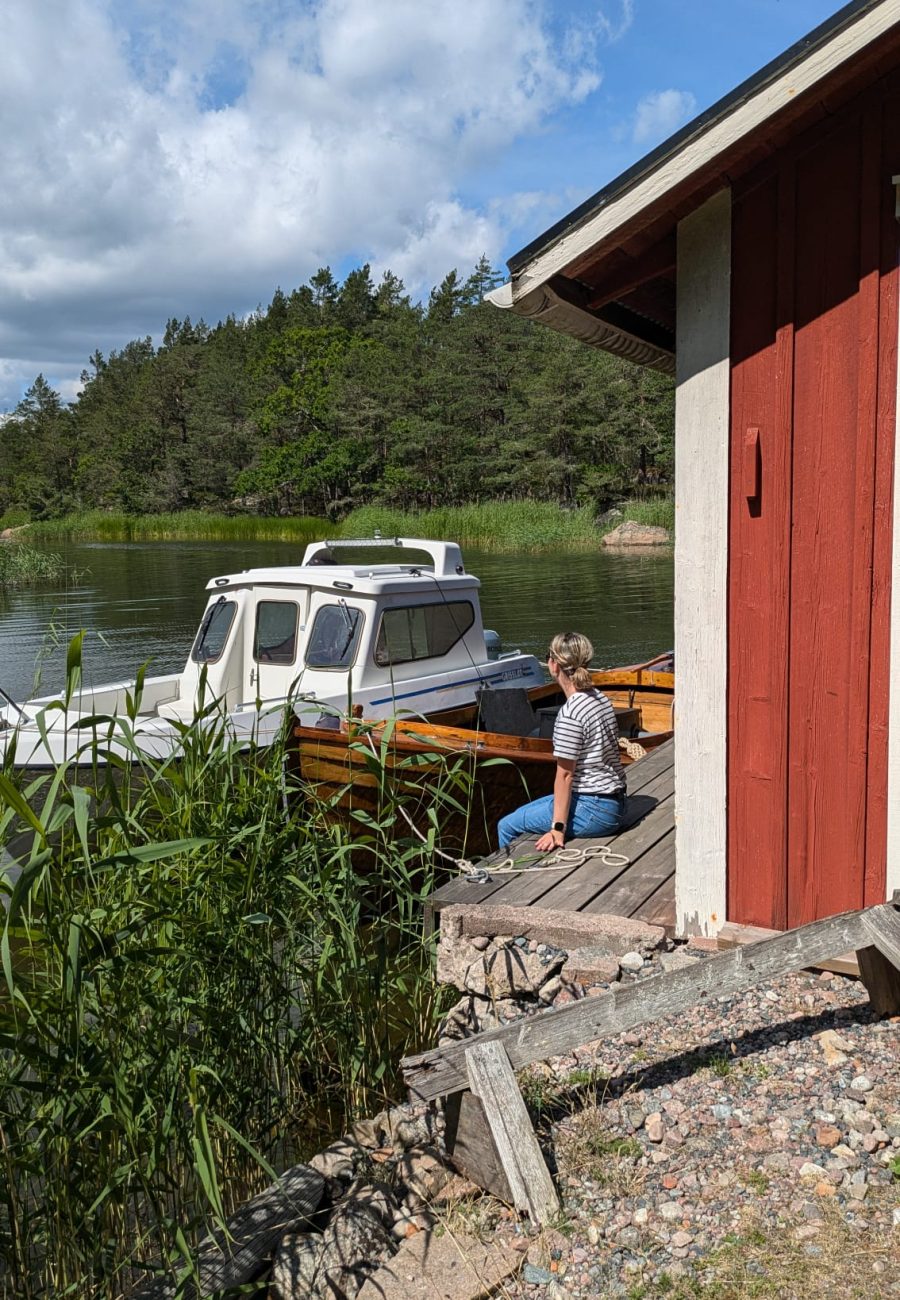 Eine Person in einem gestreiften Hemd sitzt auf einem hölzernen Steg neben einer roten Hütte und genießt Urlaub in Schweden, während sie auf ein weißes Boot auf einem ruhigen, von Bäumen und hohem Gras umgebenen See unter einem teilweise bewölkten Himmel blickt.