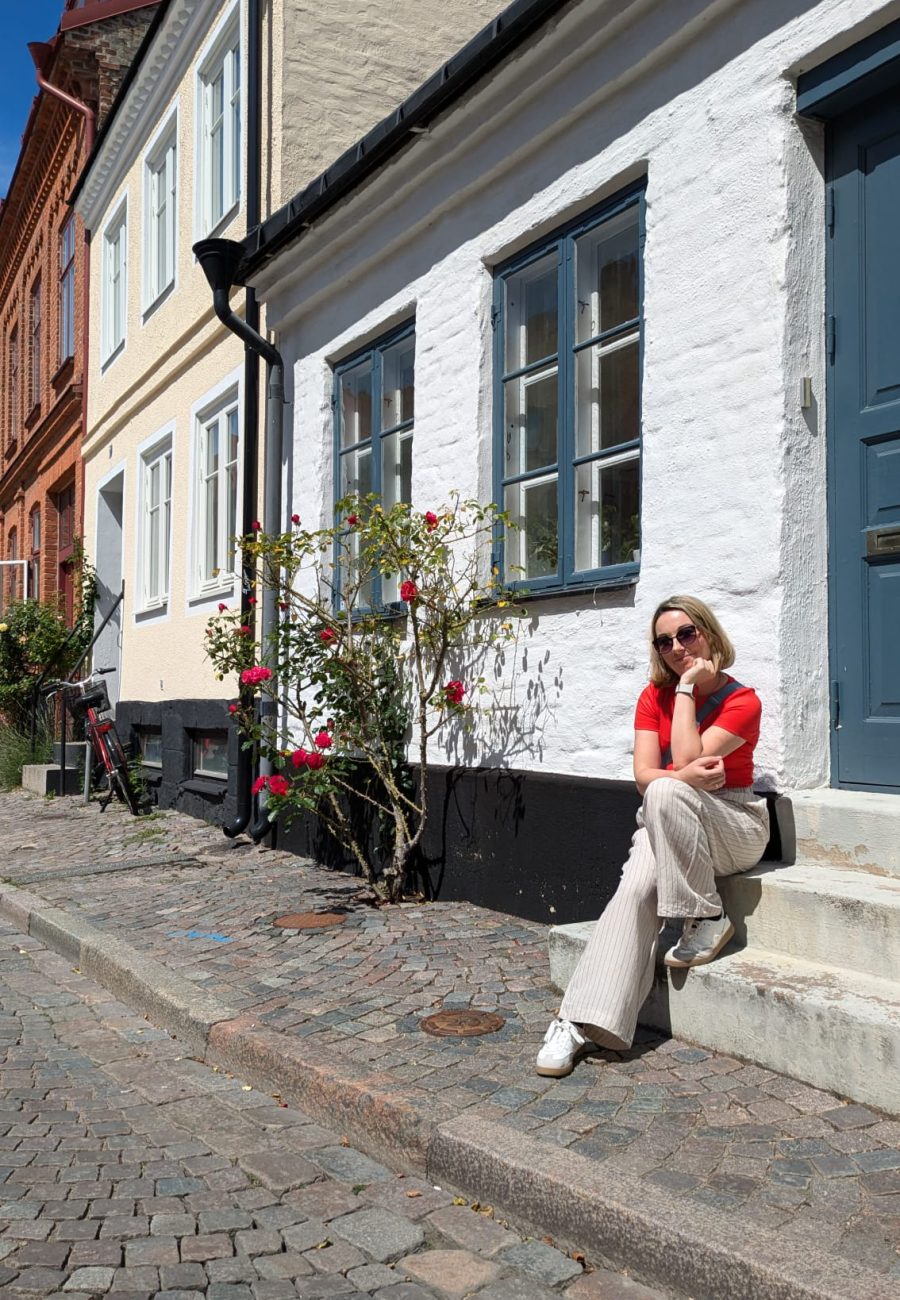Eine Frau mit Sonnenbrille sitzt auf der Türschwelle eines weißen Hauses mit blauen Türen und blühenden Rosensträuchern und genießt die sonnige Kopfsteinpflasterstraße mit ihren bunten Häusern - perfekt für einen idyllischen Urlaub in Schweden.