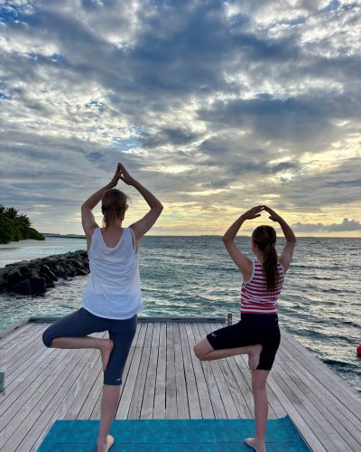 Zwei Personen in einer Yoga-Pose auf einem Holzsteg am Meer auf den Malediven bei Sonnenaufgang.